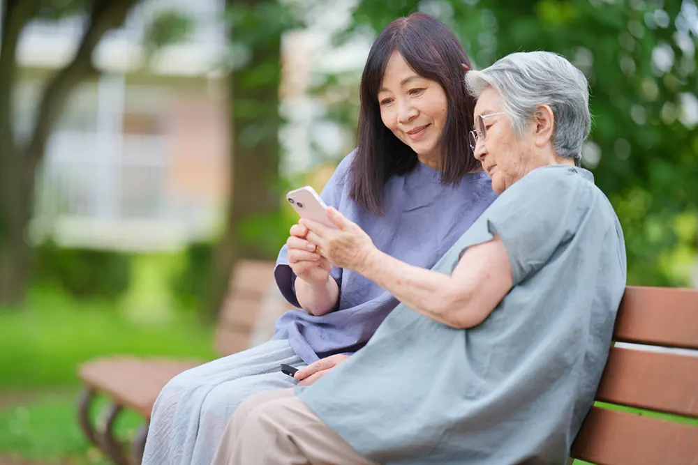 ベンチで高齢女性にスマートフォンの画面を見せる若い女性